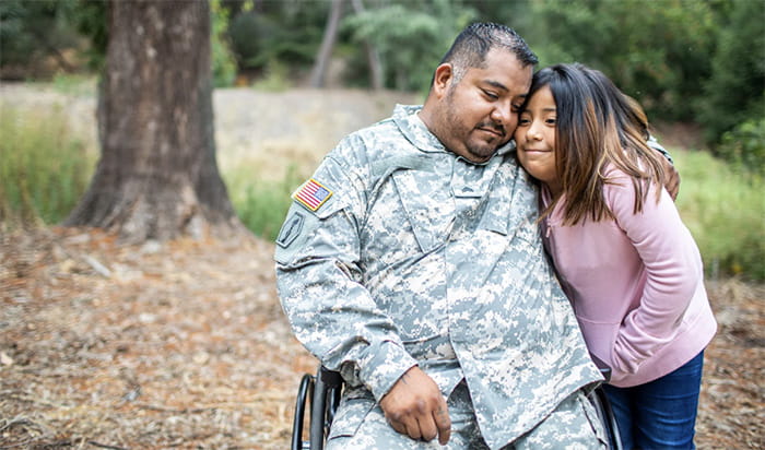 A man wearing military camouflage and seated in a wheelchair embraces a small girl with his left arm.