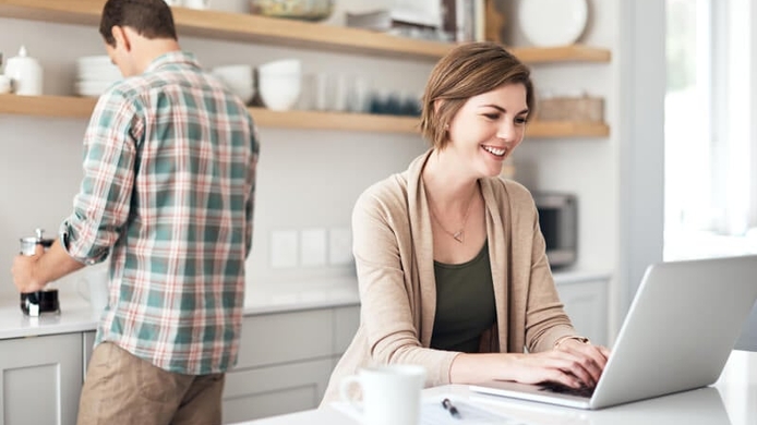 female at a laptop in the kitchen while male spouse prepares something behind her