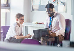 A male secondary school student sitting in an office in the school, having a meeting with his teacher. The teacher is showing him something on his laptop while they both smile.