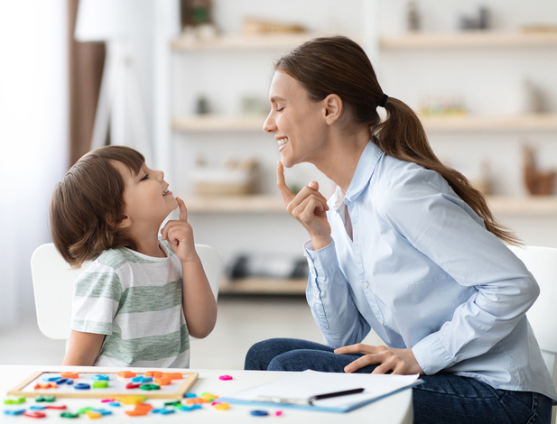 Speech training for kids. Professional woman specialist training with little boy at cabinet, teaching him right articulation exercises, side view
