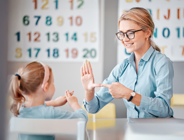 Smiling caucasian teacher wearing spectacles communicating with deaf girl student with hand sign language in a classroom at school. Sign language teacher in a tutoring class for a cute female child with hearing disability